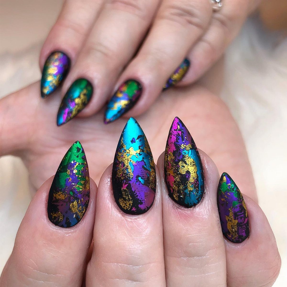 Stiletto nails with purple, green, and gold marbled metallic nail art.