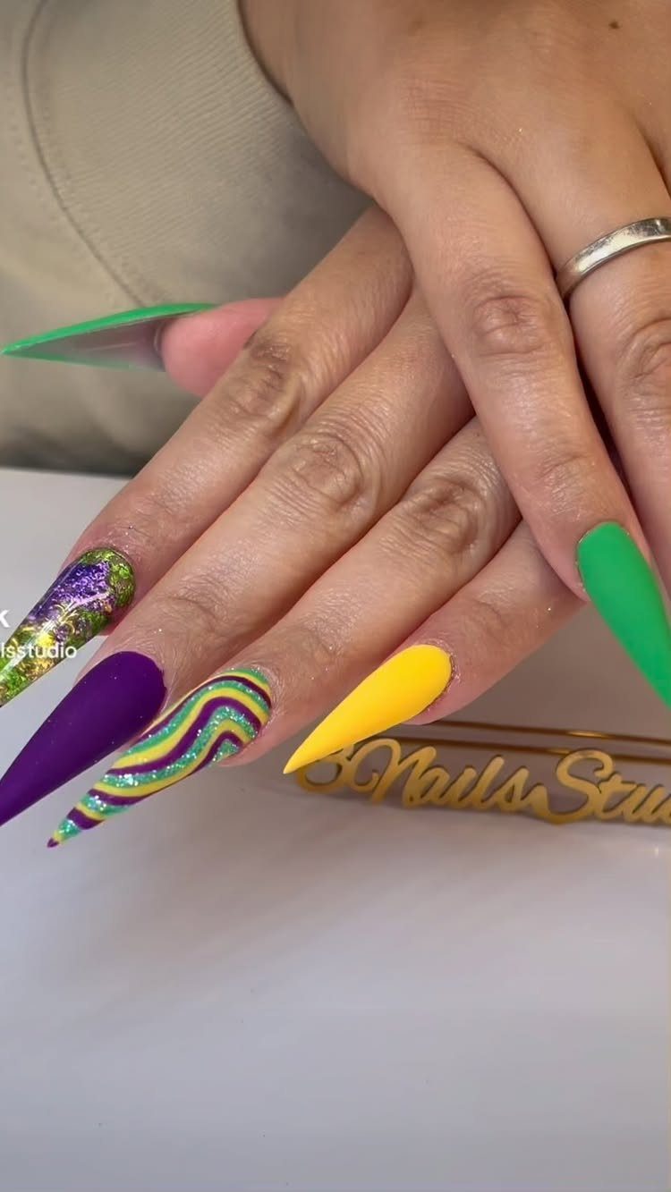 Stiletto nails painted in purple, green, and yellow with glitter and swirls.