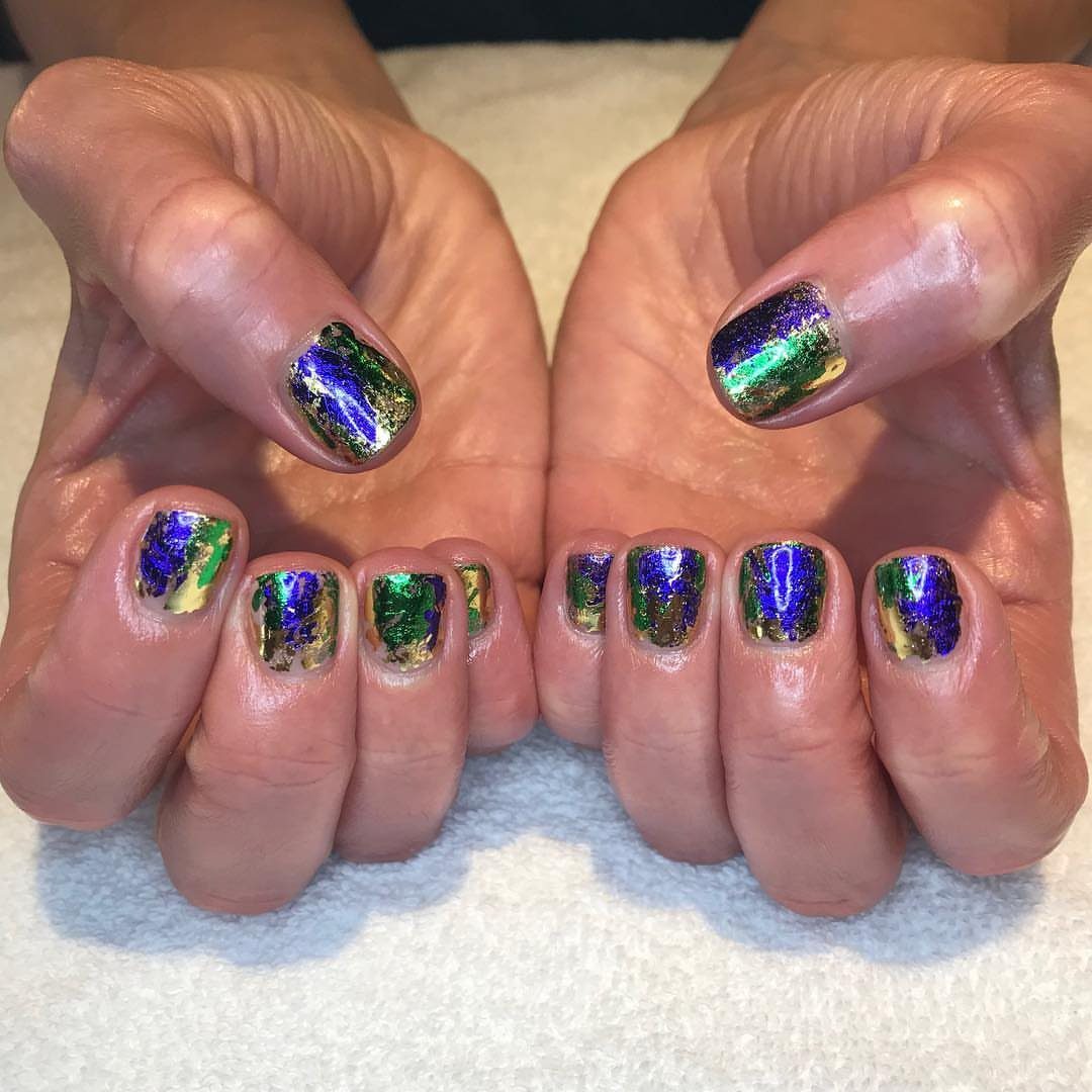 Short nails decorated with shimmering purple, green, and gold metallic carnival foil.