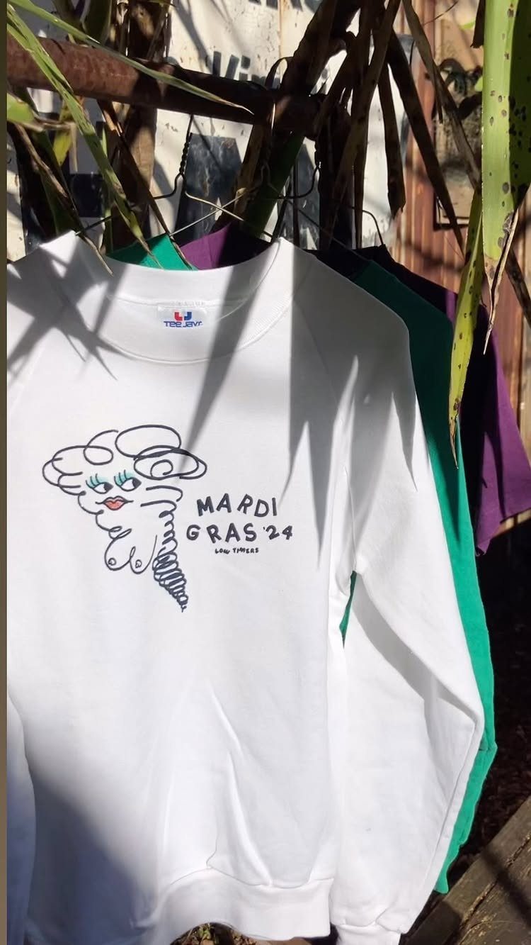 White Mardi Gras 24 sweatshirt hanging outdoors with colorful shirts behind it.