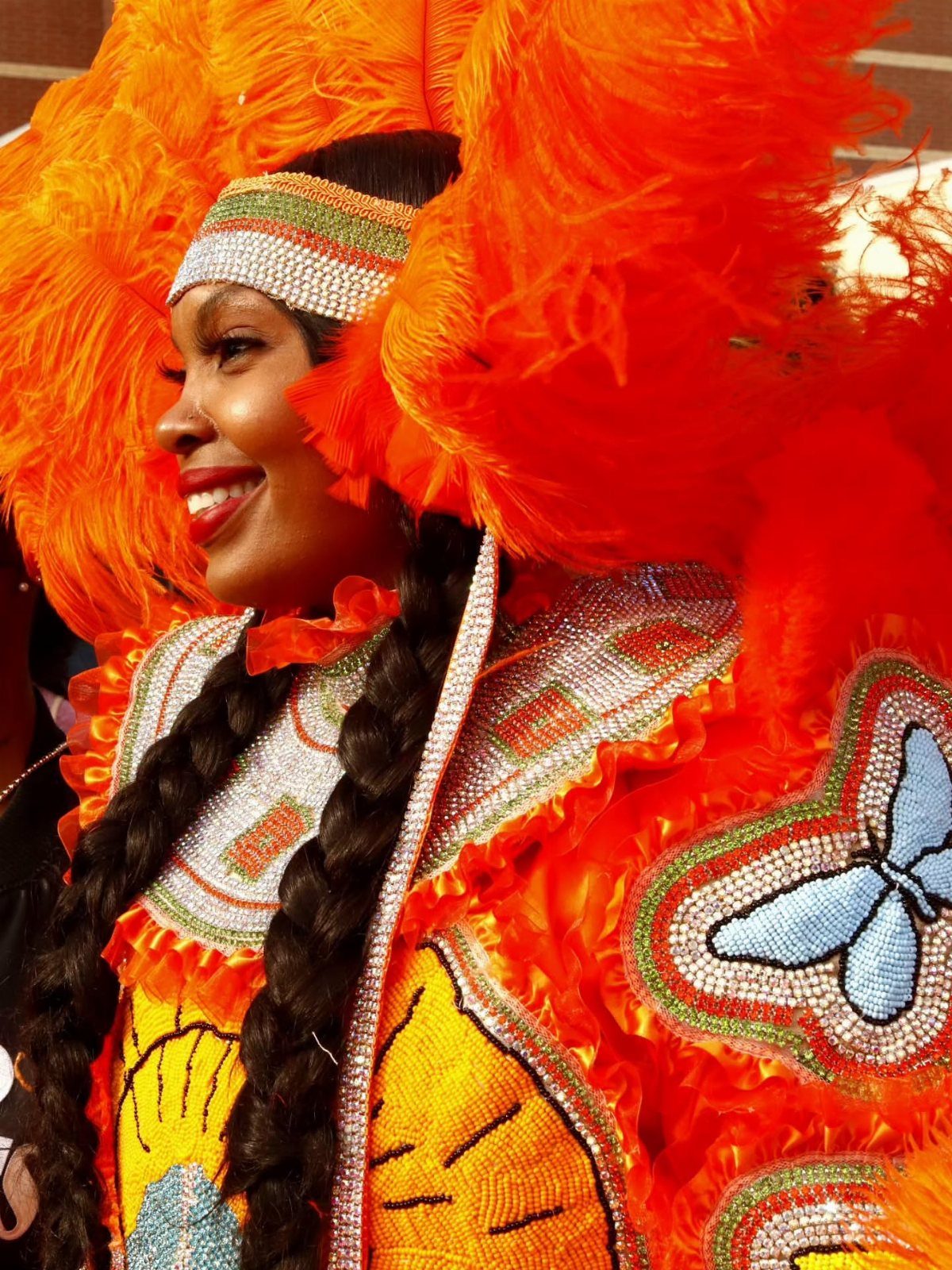 Woman in an elaborate orange feathered headdress and sequined costume smiling outdoors.