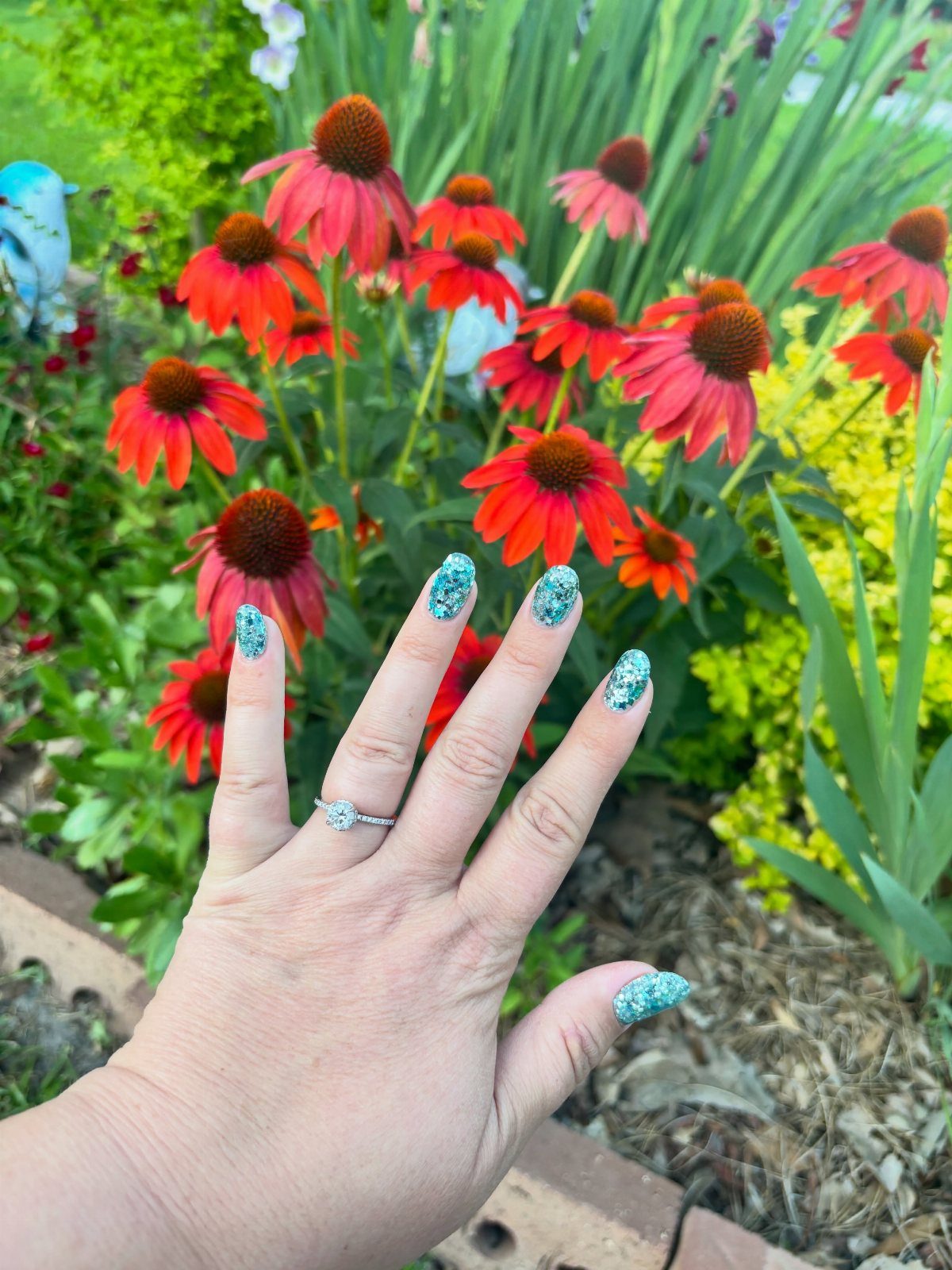 Hand with blue glitter nails posing in front of red coneflowers in a garden