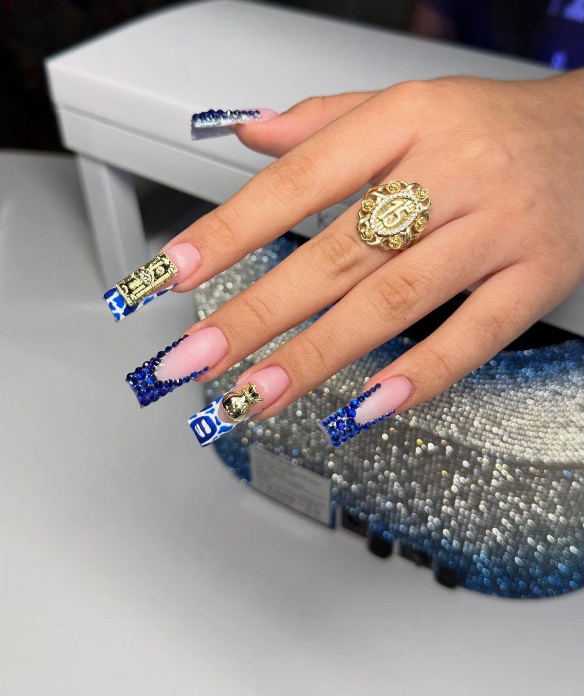 Long acrylic nails with blue glitter tips and gold charms on a hand.