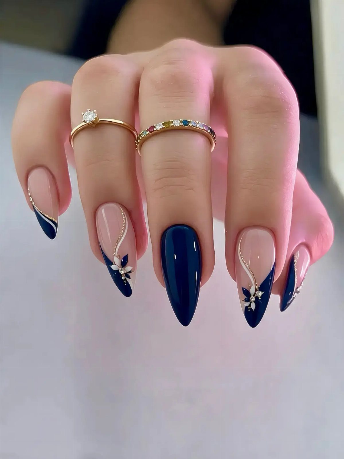 Navy blue and nude almond nails with white floral accents and gold rings.