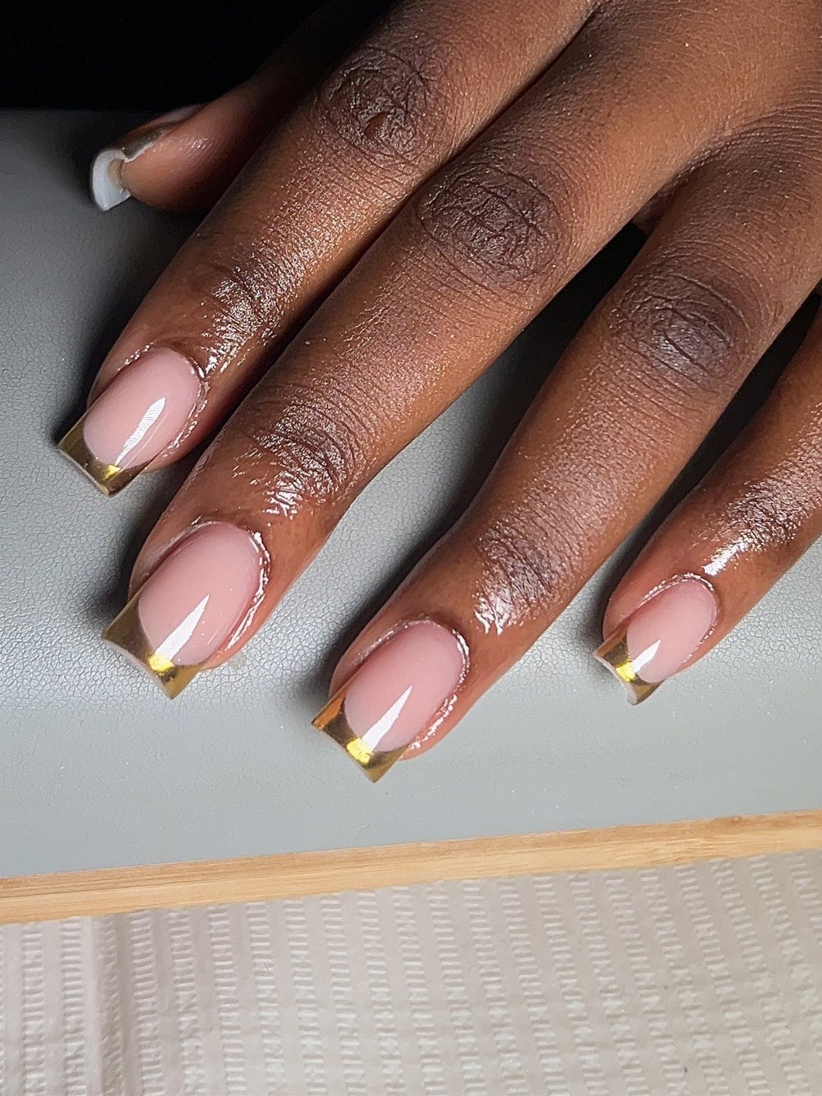 Nude oval nails with metallic gold French tips on a dark-toned hand.