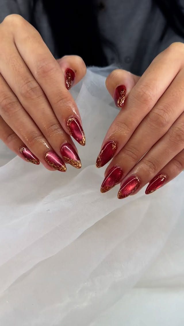 Deep red almond-shaped nails with gold glitter accents on white fabric.
