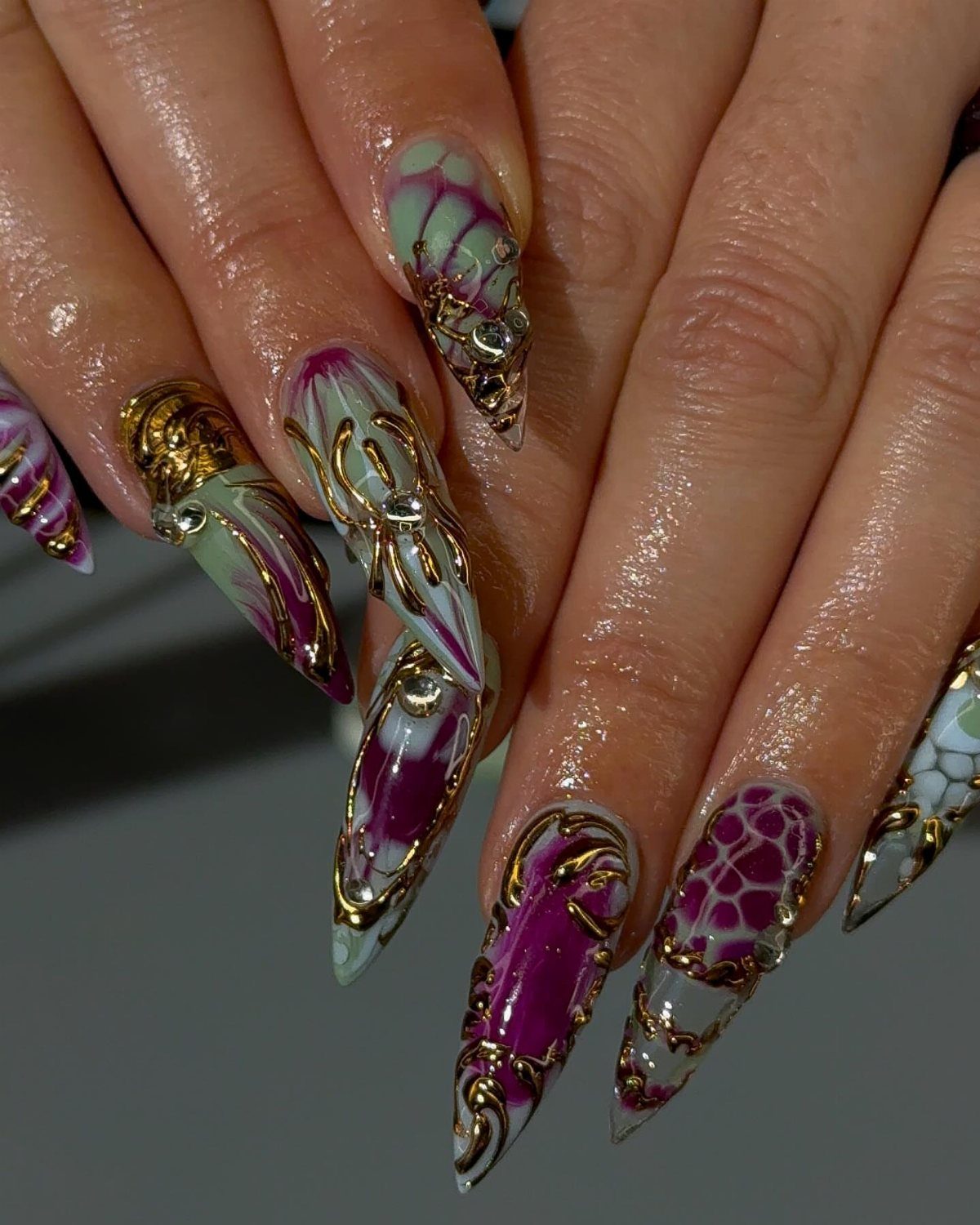 Stiletto nails with purple, green, and gold marbled patterns and metallic gold accents.