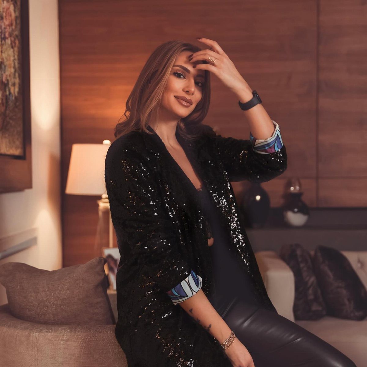 Woman wearing a black sequined blazer and leather trousers sitting on a sofa.