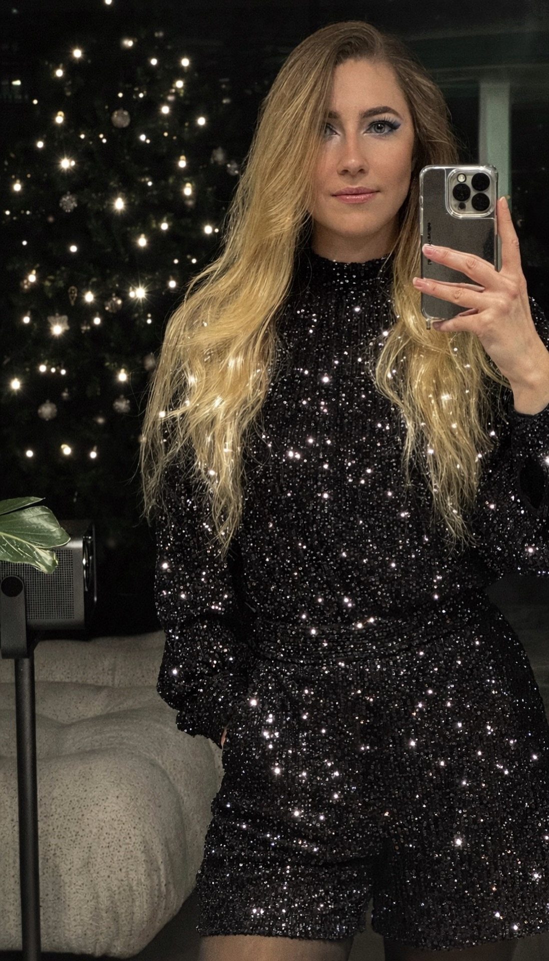 Woman in a black sequined top and matching shorts posing by a Christmas tree.