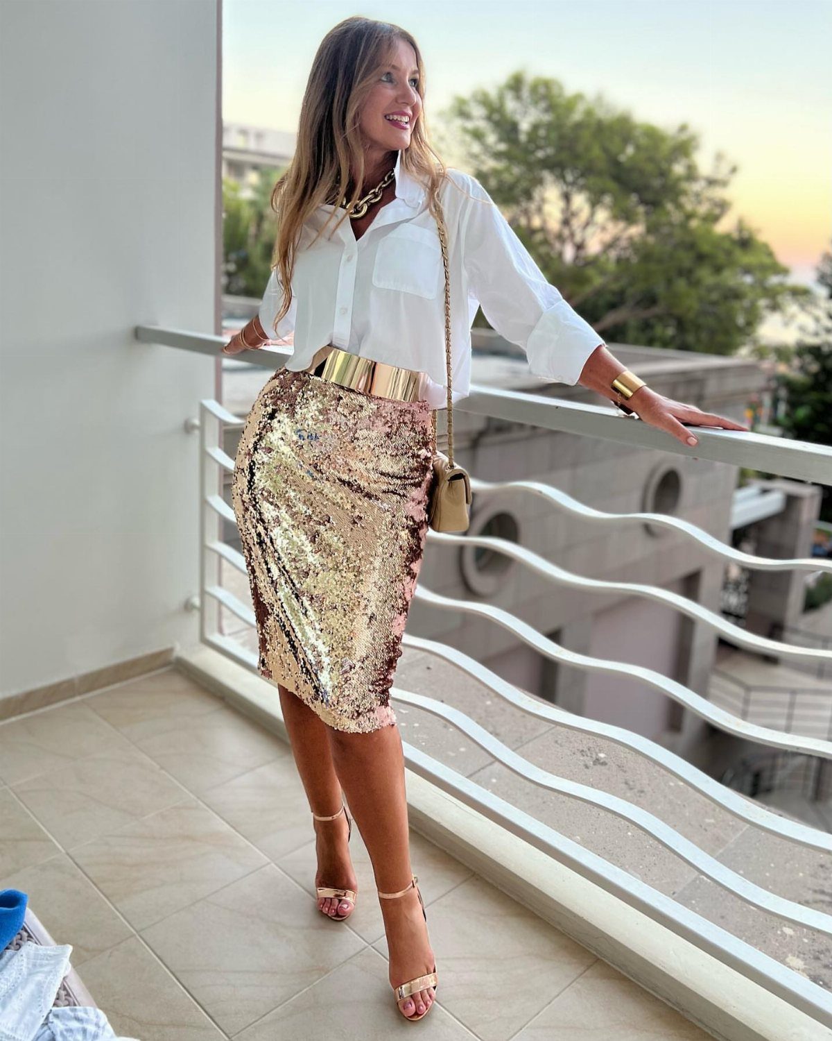 Woman in a white button-down shirt and gold sequined midi skirt on a balcony.