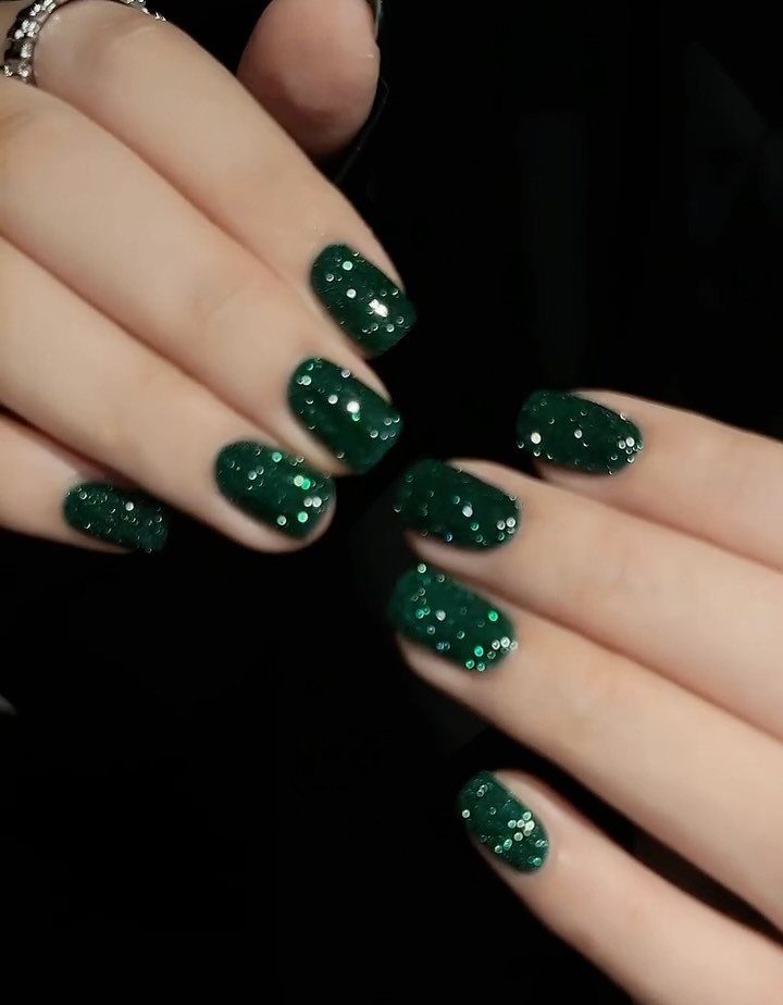 Deep emerald green glitter nails on long oval fingertips against a black background.