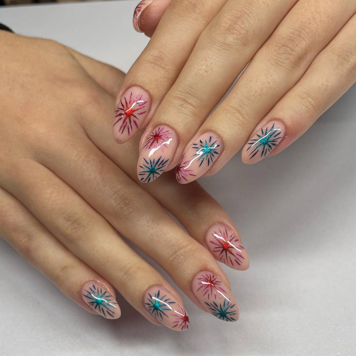 Almond-shaped nails with colorful red and blue firework starburst designs on sheer base.