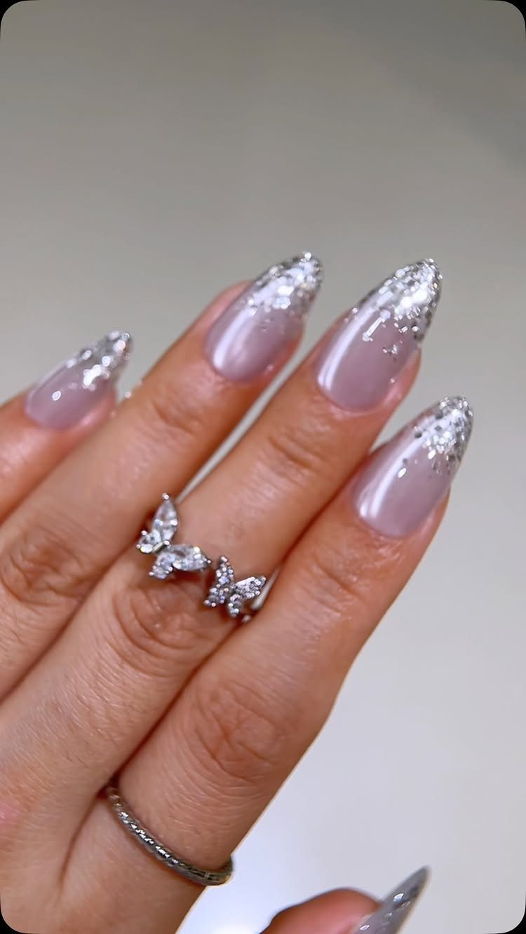 Almond-shaped nails with light pink base and silver glitter ombré tips and rings.