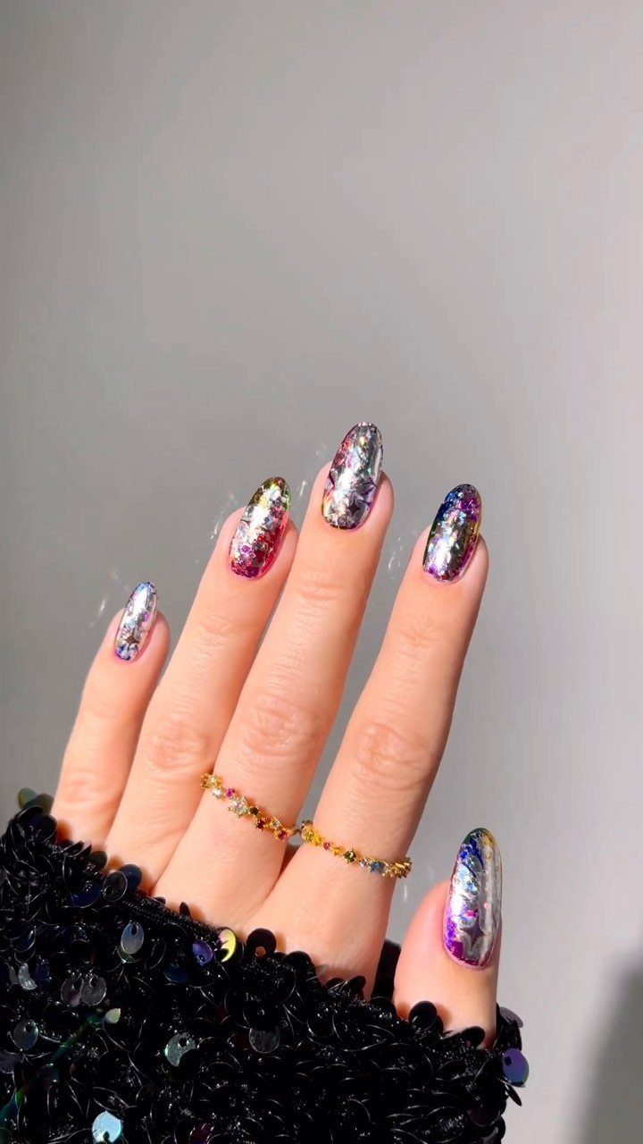 Holographic nails with color-shifting iridescent polish on a hand with gold rings.