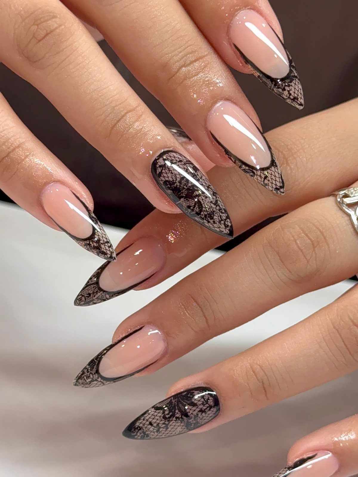Almond nails with sheer nude bases and intricate black lace pattern tips.
