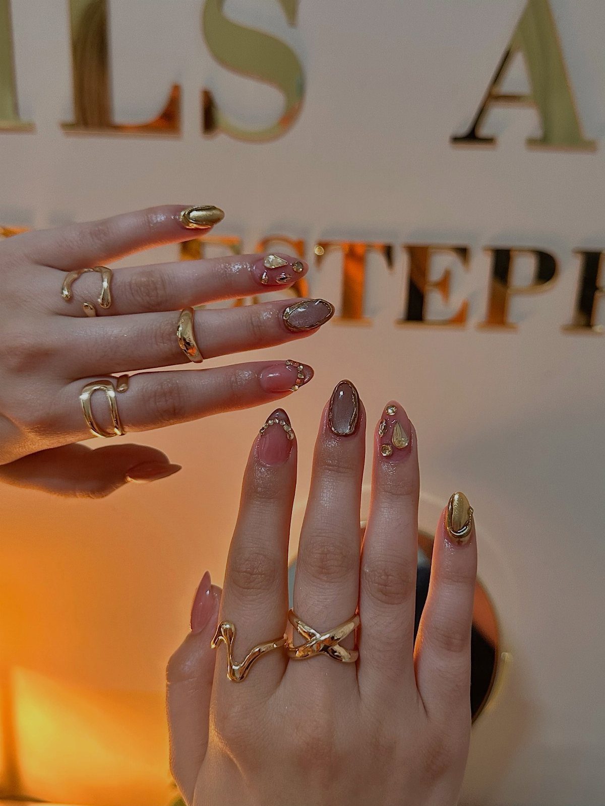 Manicured nails with mismatched gold, brown, and pink designs and gold rings.