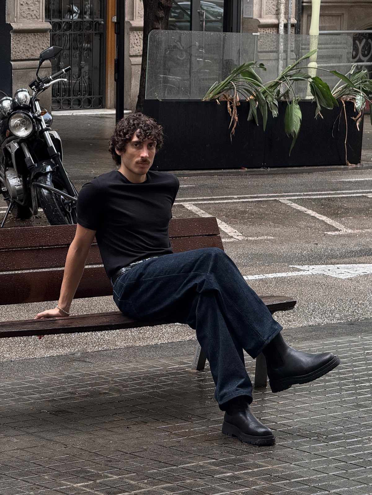 Man sitting on a bench wearing a black t-shirt, dark jeans, and black boots.