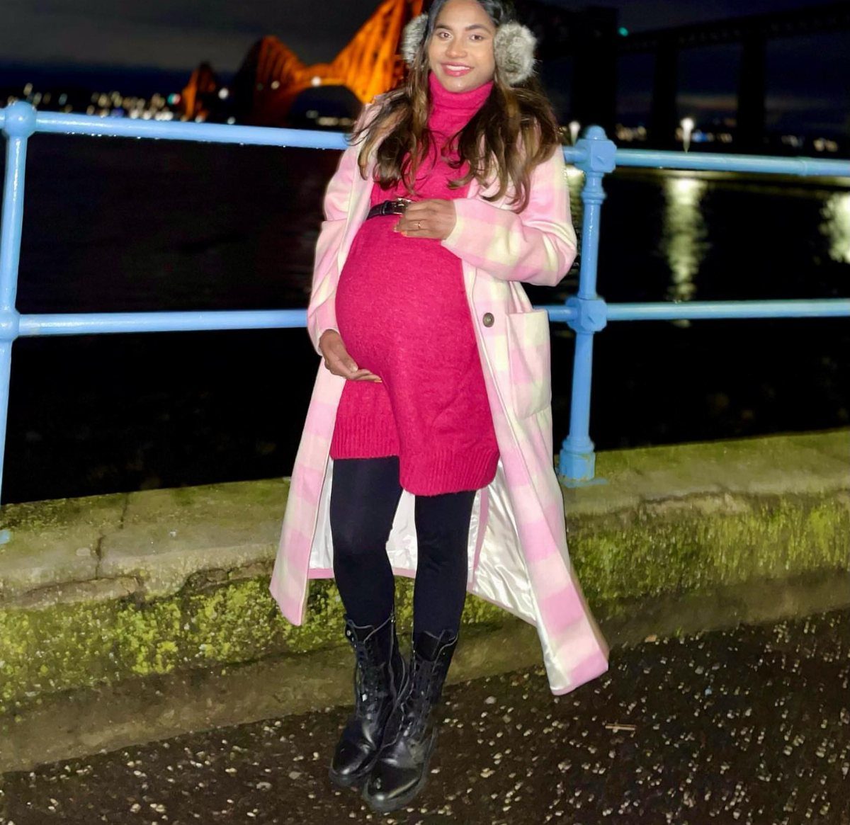 Pregnant woman in a pink checkered statement coat and pink sweater dress outdoors.
