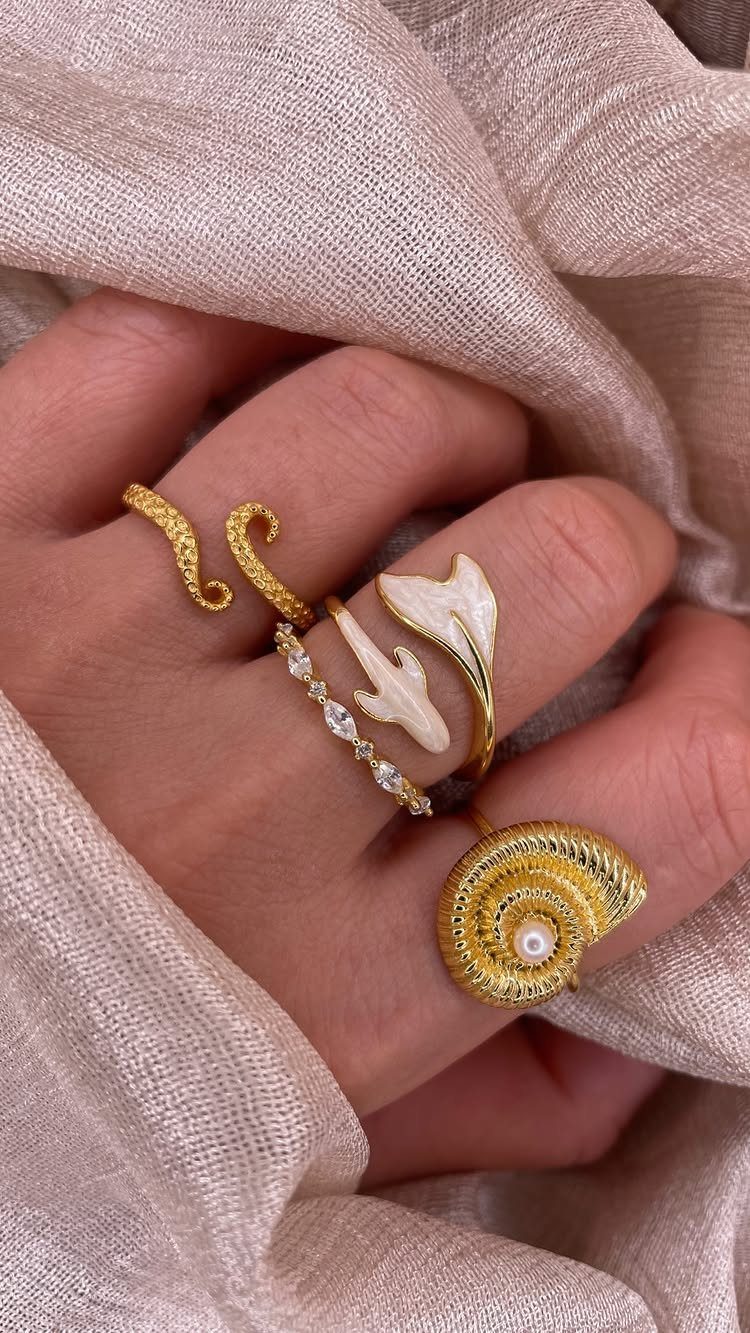 Gold-toned beach-themed rings on a hand resting against beige textured fabric.