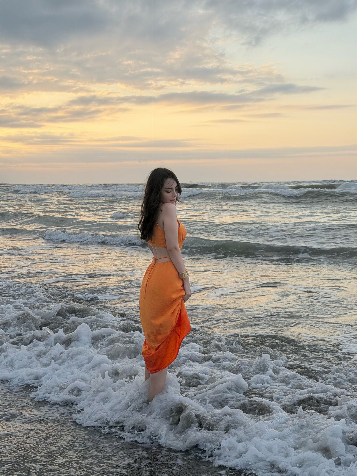 Woman in orange two-piece outfit standing in ocean surf at sunset beach