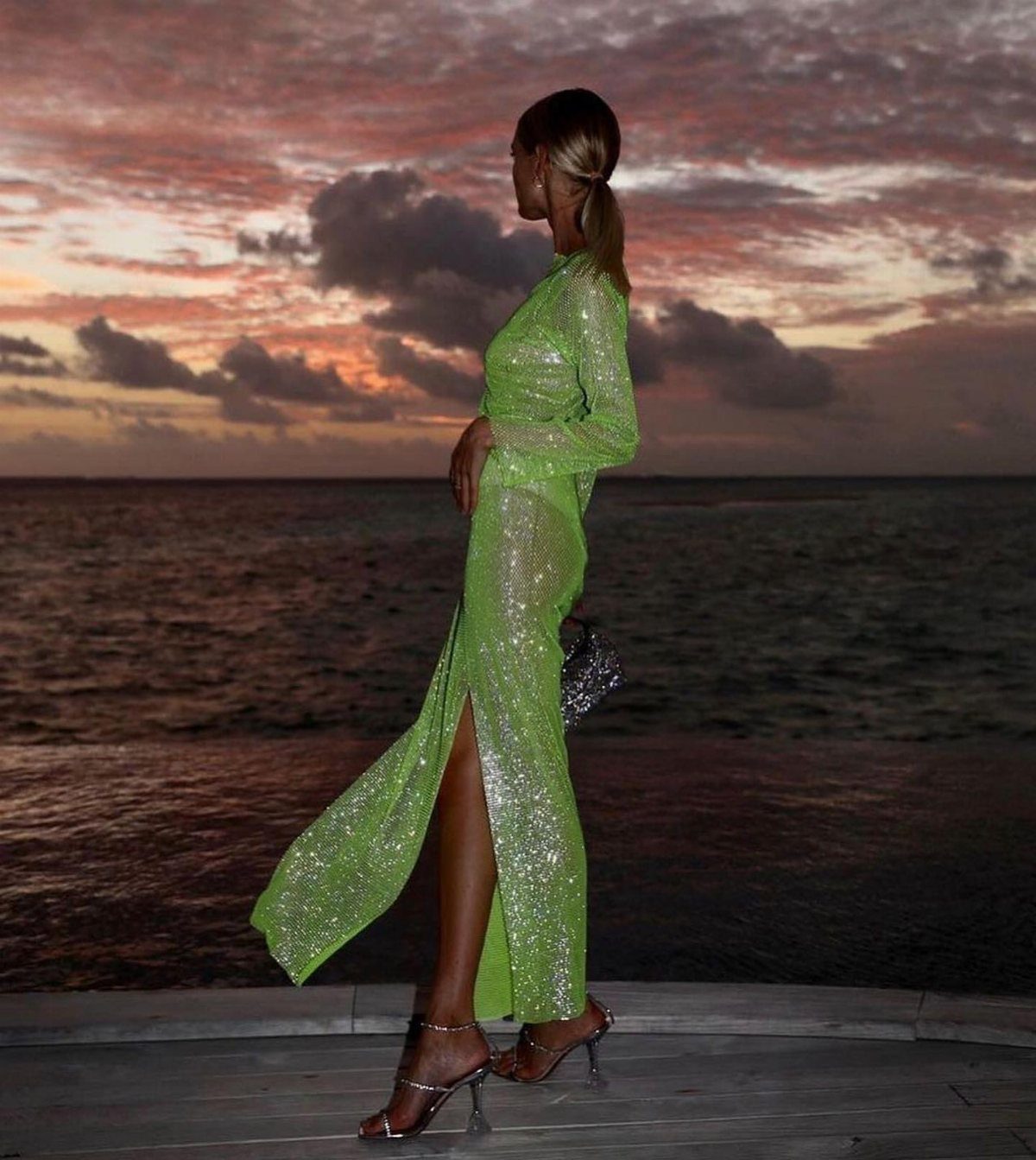 Woman in a lime green sequined gown standing on a beach deck at sunset