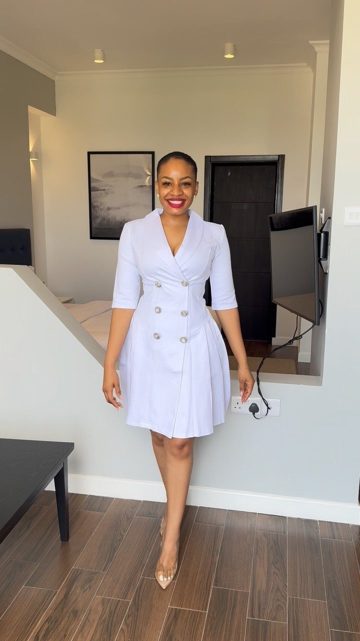 Woman wearing a white double-breasted blazer dress and clear heels in a modern room.