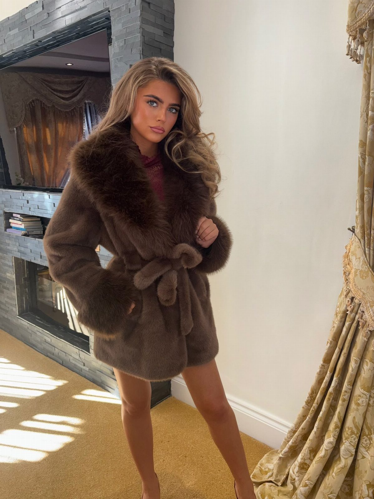 Woman wearing a brown belted faux fur coat standing in a living room.