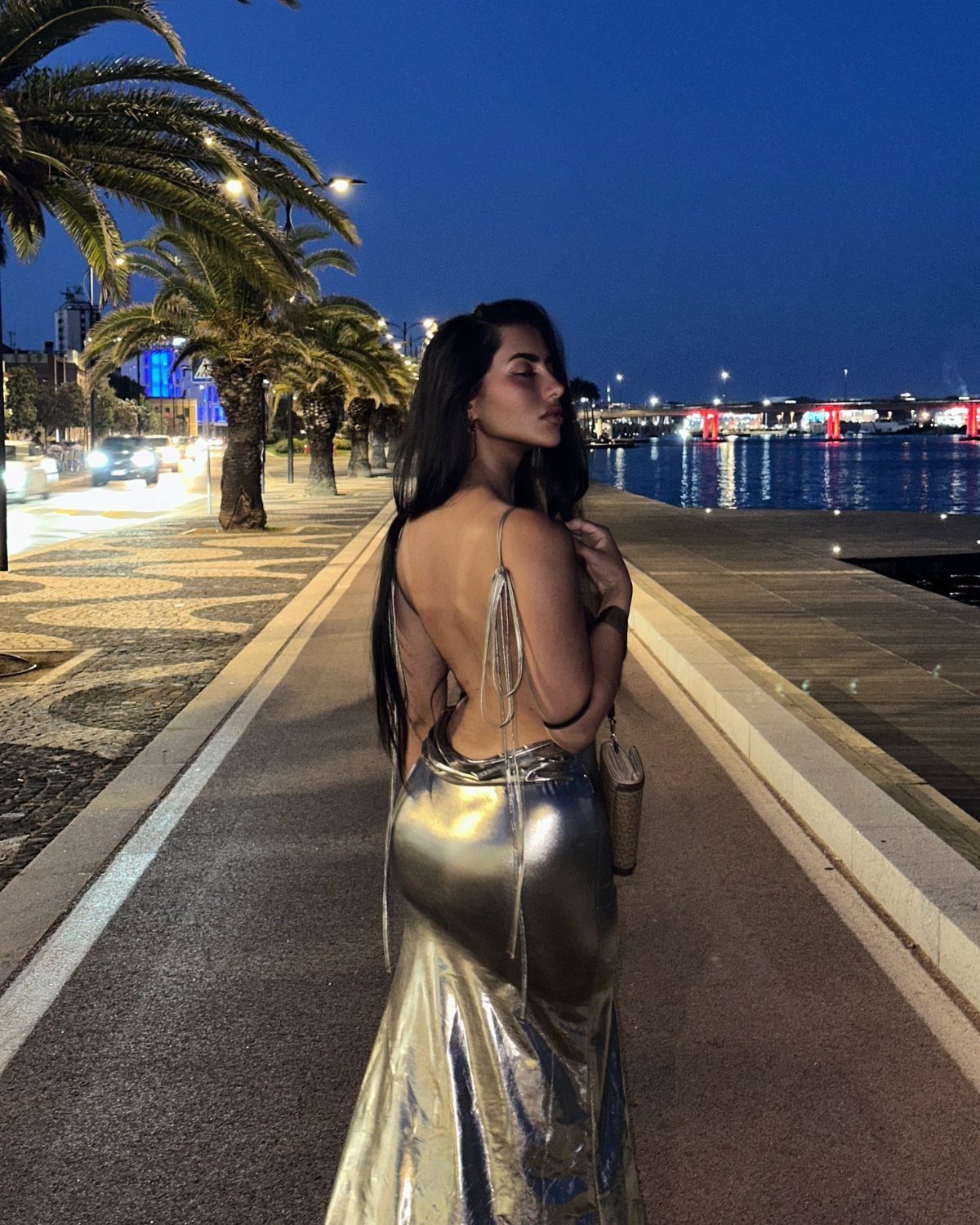 Woman in a backless metallic silver gown standing on a waterfront at night.