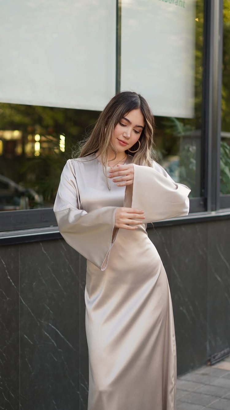 Woman in a champagne satin long-sleeved maxi dress standing by a window.