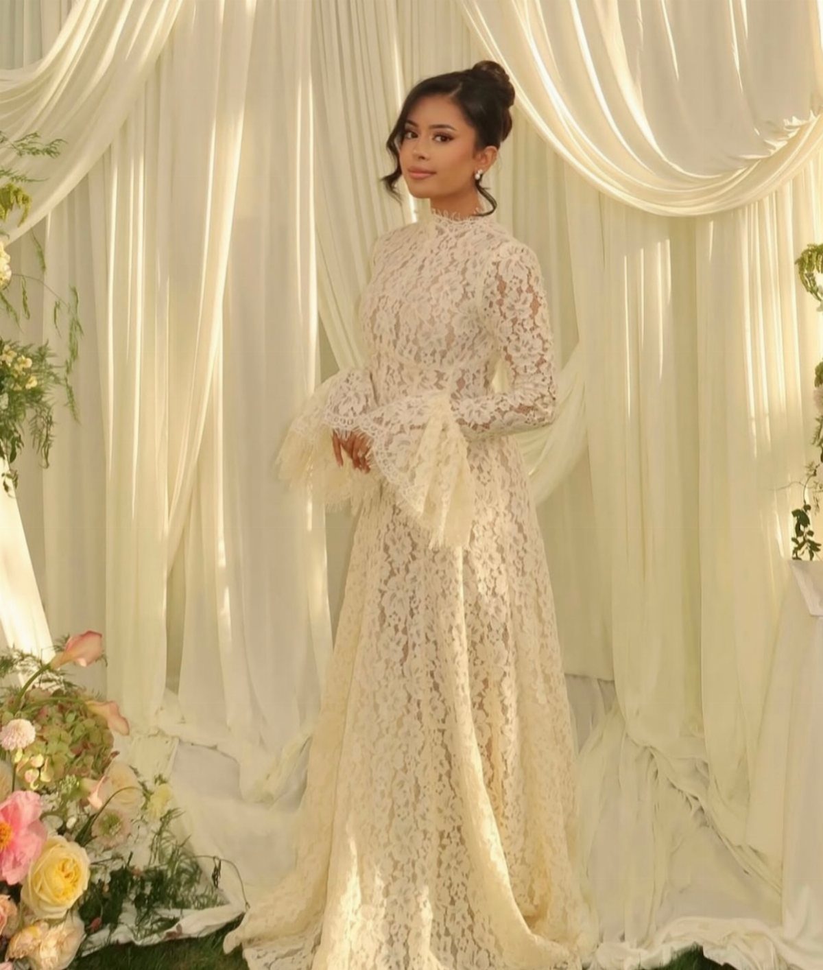 Woman in a cream lace A-line maxi dress with flutter sleeves against white drapes.