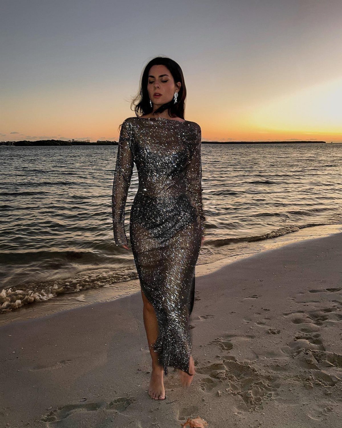 Woman in a long-sleeved silver sequined mesh maxi dress standing on a beach.