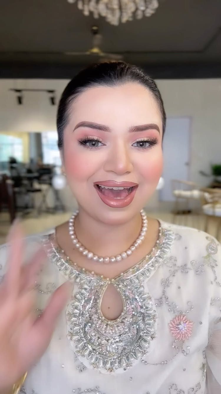 Woman with pink eyeshadow and glossy lips wearing a white embroidered dress and pearls.