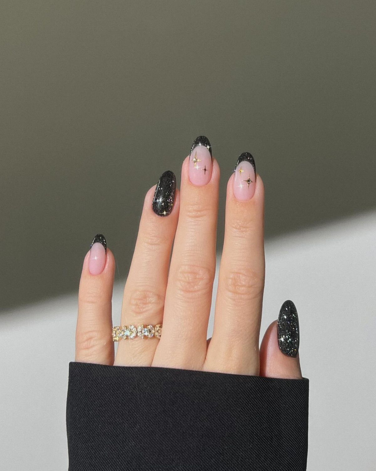 Hand with black glitter and nude nails featuring gold star accents and French tips.