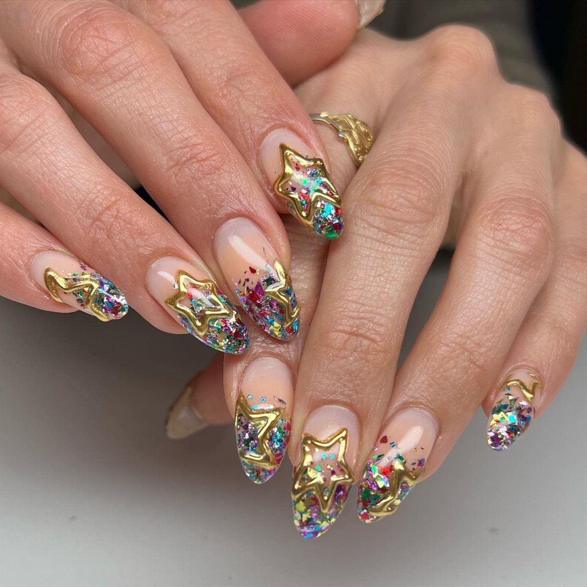 Nude almond nails decorated with colorful glitter confetti and 3D gold stars.