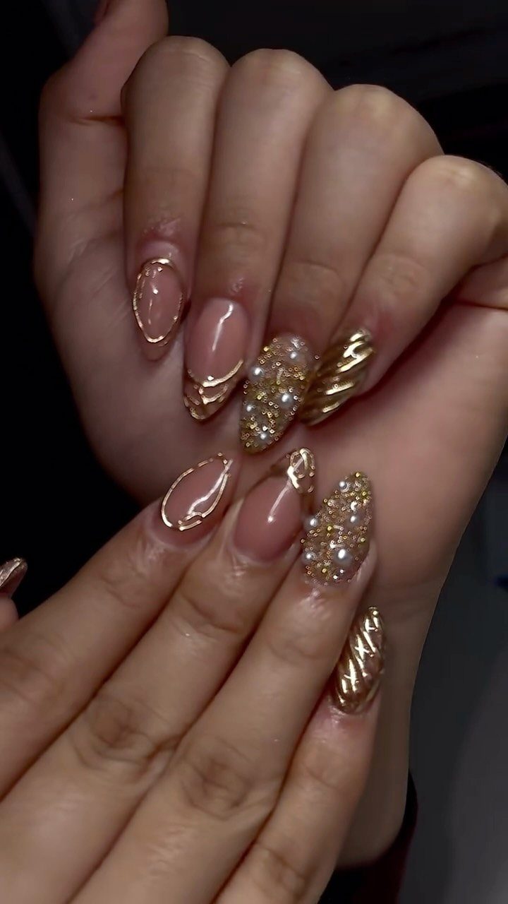 Long almond nails with metallic gold swirls, gold glitter, and pearl accents.