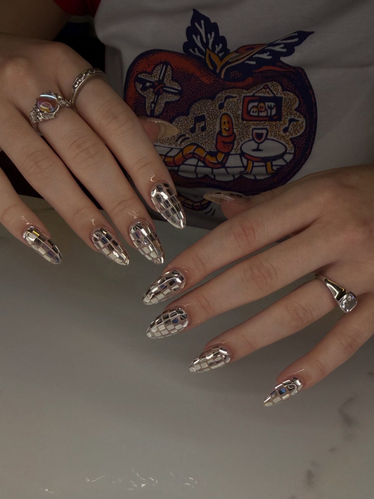 Long almond nails with silver mirrored mosaic disco ball patterns and silver rings.