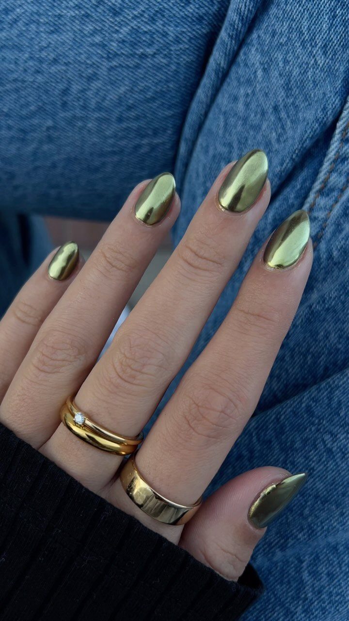Almond-shaped nails with metallic gold chrome polish and gold rings on a hand.
