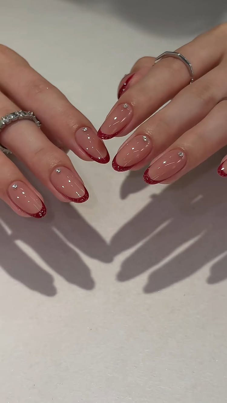 Almond-shaped nails with red glitter French tips and small rhinestone accents on nude base.
