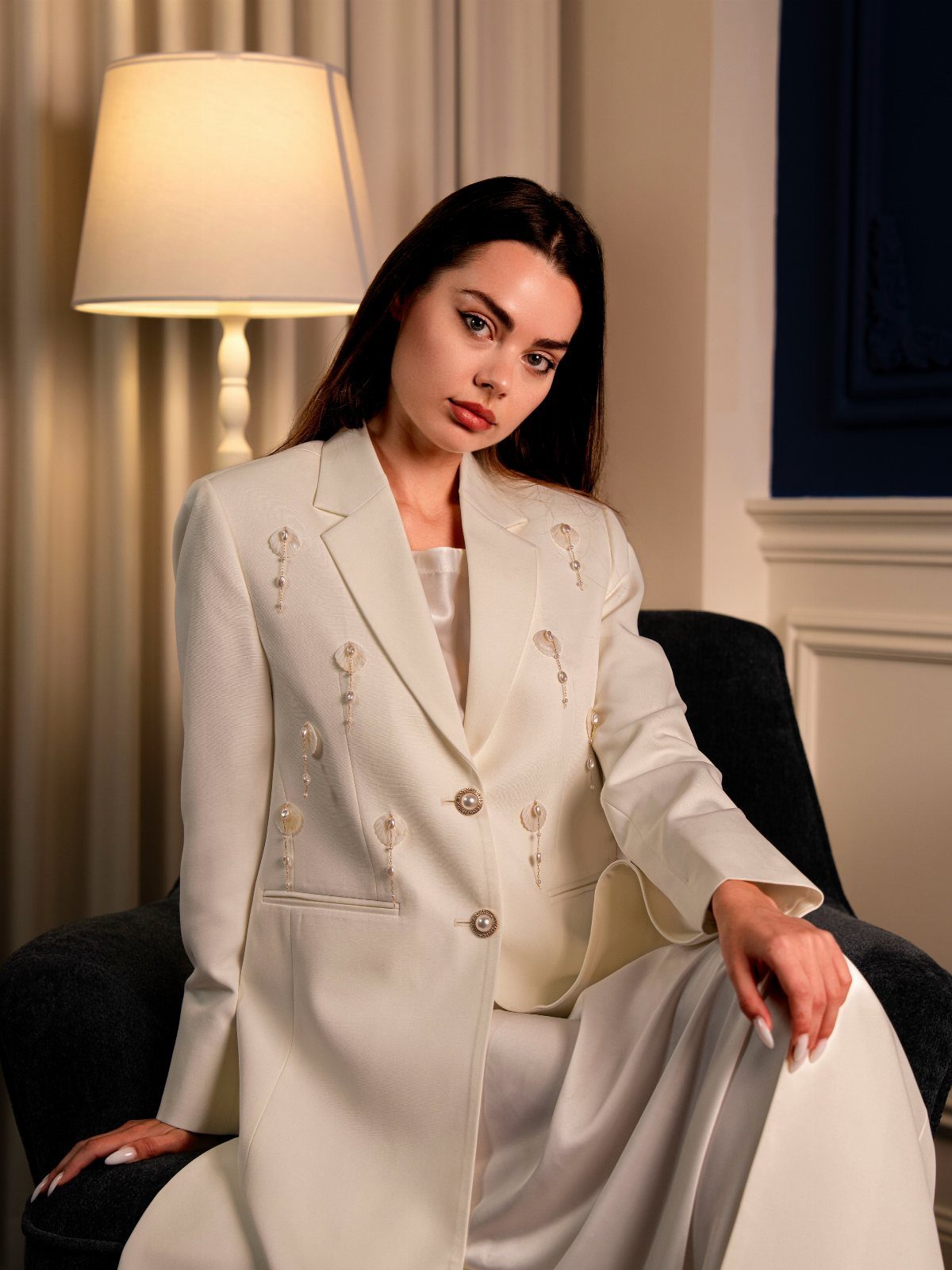 Woman wearing an oversized white blazer and trousers sitting in a grey armchair