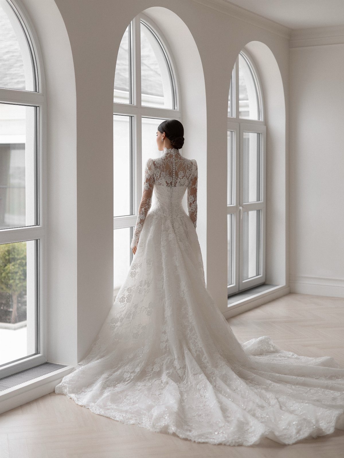 Woman in a white long-sleeved lace wedding gown standing by arched windows.