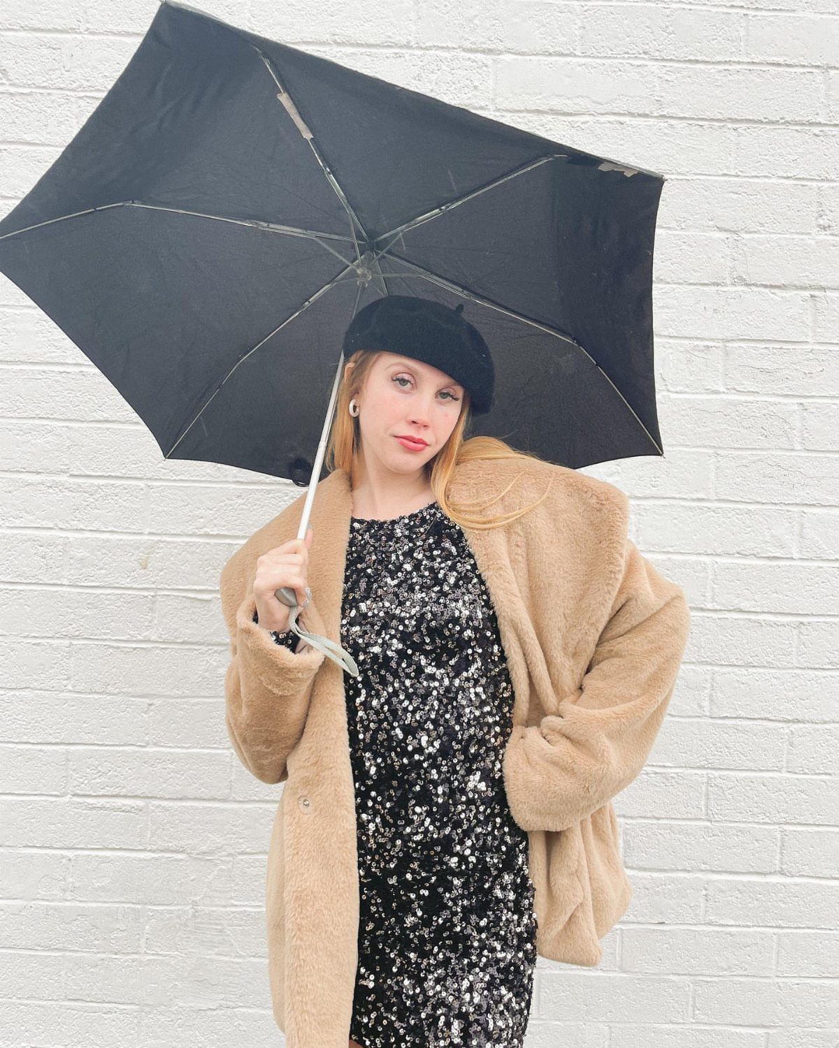 Woman in a sequined dress, tan teddy coat, and beret holding a black umbrella.