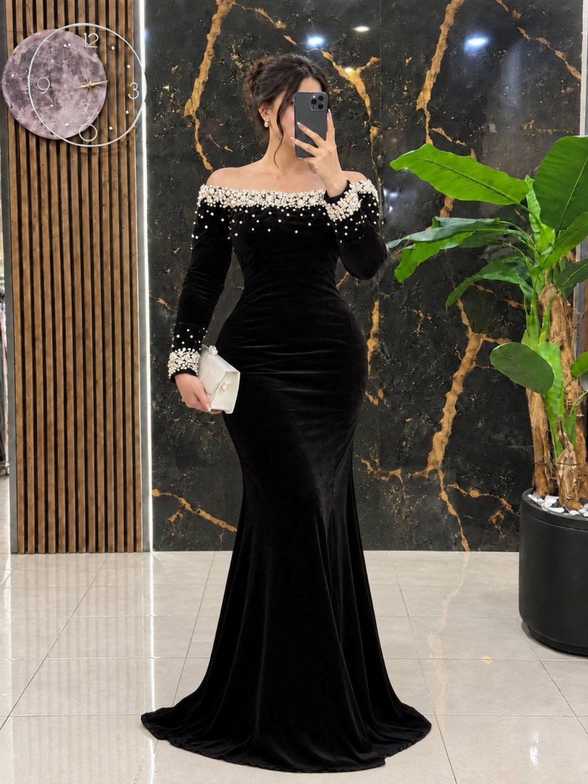 Woman in a black velvet mermaid dress with pearl details and white clutch.