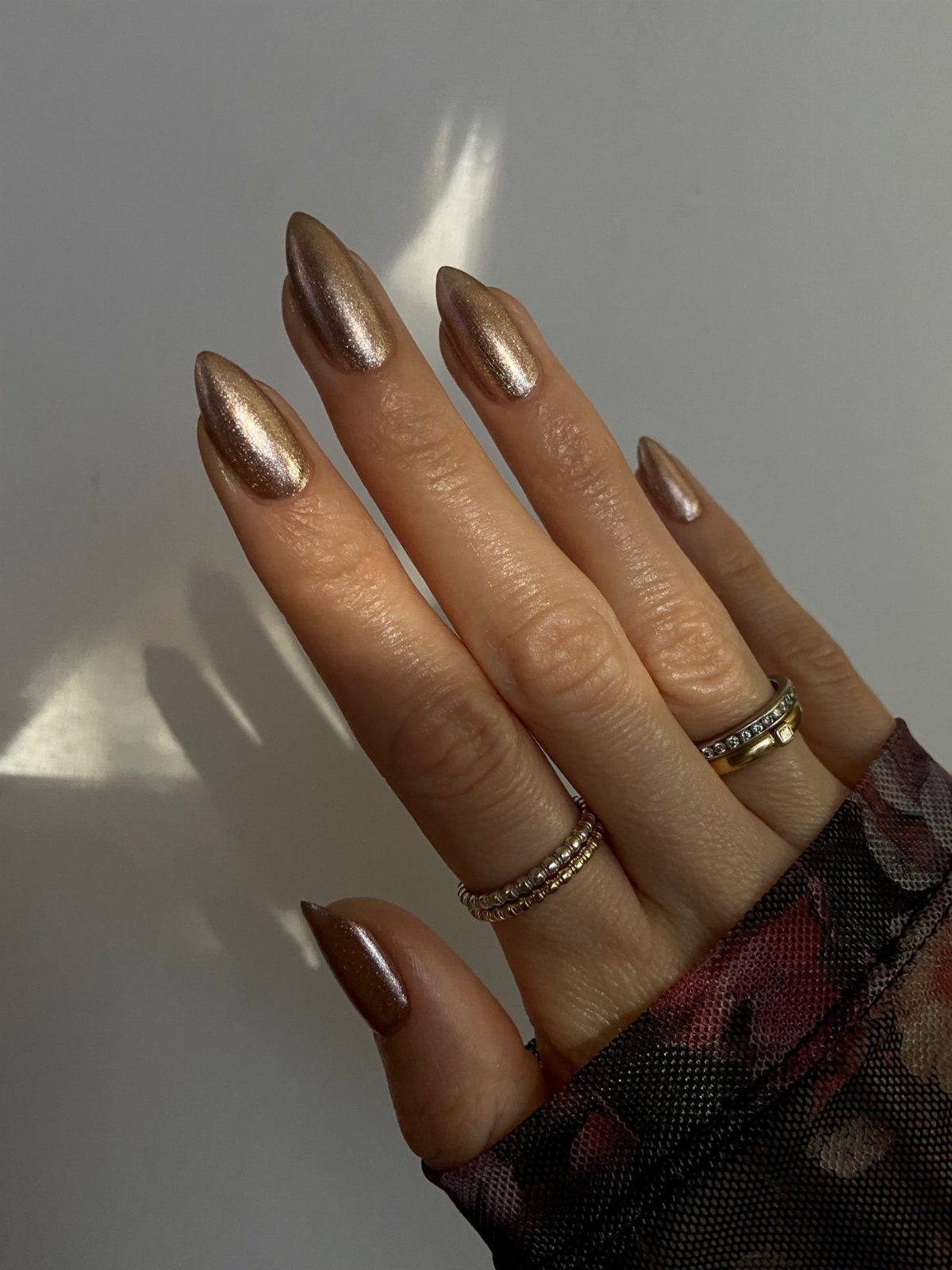 Almond-shaped nails with shimmering metallic gold-bronze polish and gold rings on fingers.