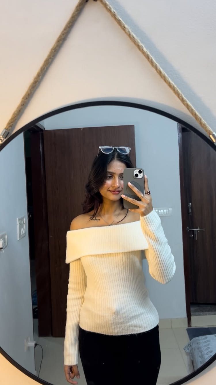 Woman in a white off-the-shoulder ribbed top and black pants taking a mirror selfie.