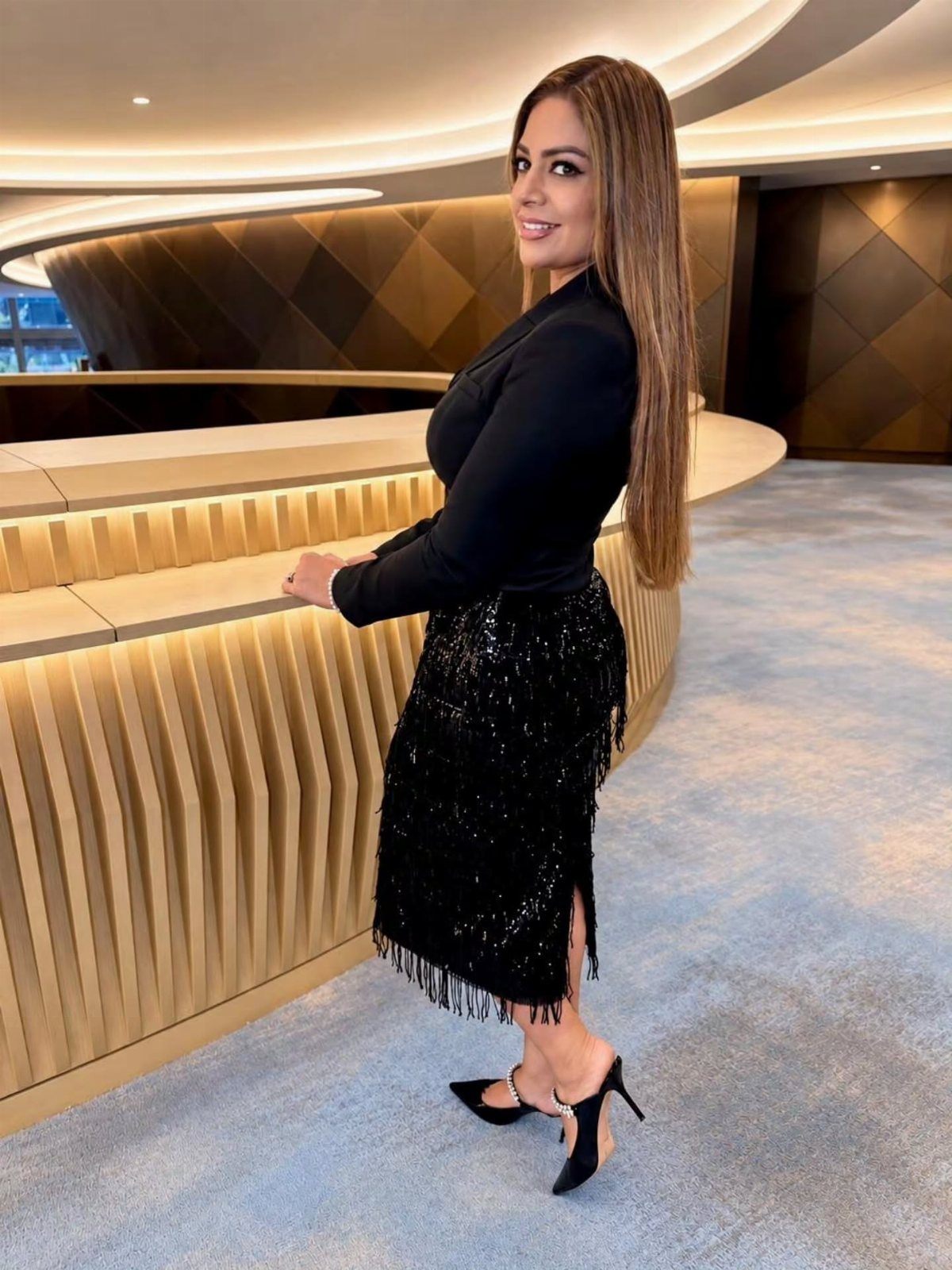 Woman in black blazer and sequined skirt posing in a modern gold interior.