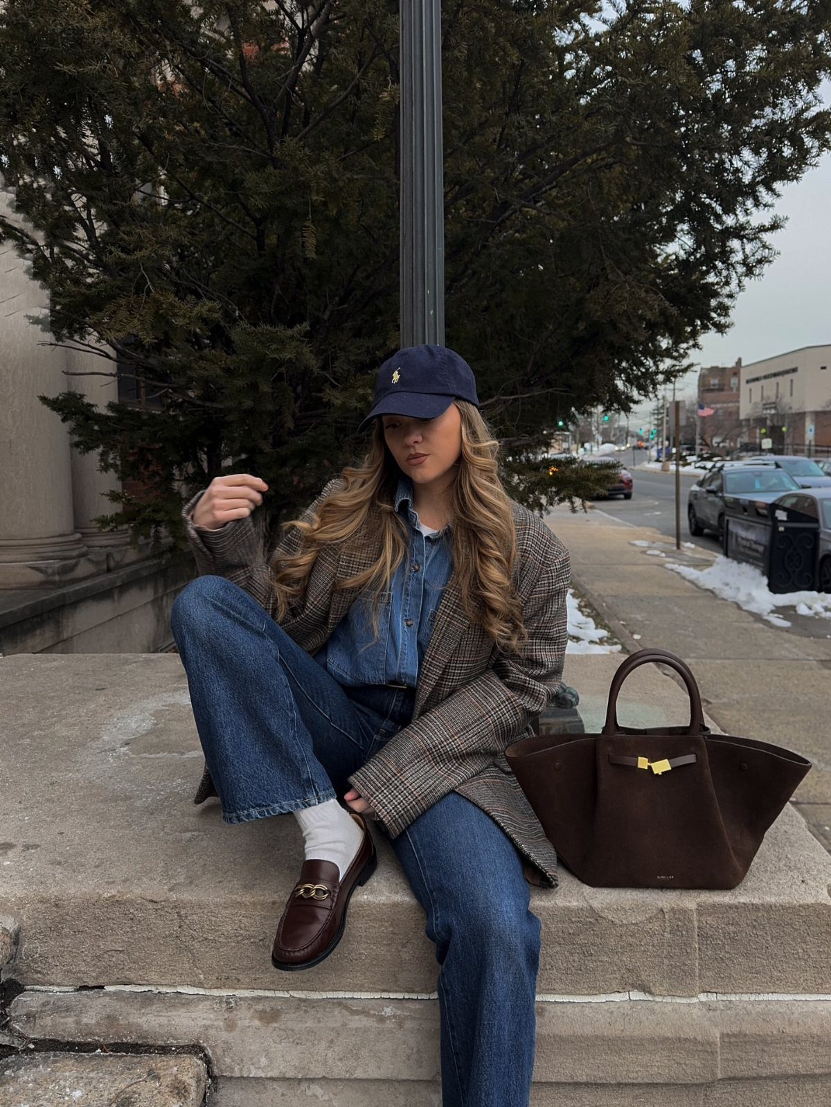 Woman in plaid blazer and denim sitting on stone steps with brown handbag