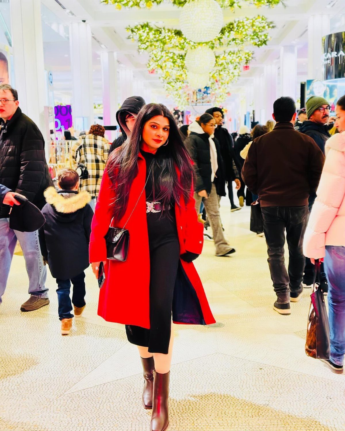 Woman in a red coat and black outfit standing in a crowded mall.