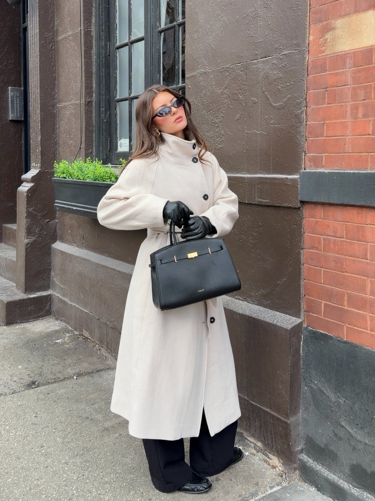 Woman in a long cream coat and black trousers posing on a city street