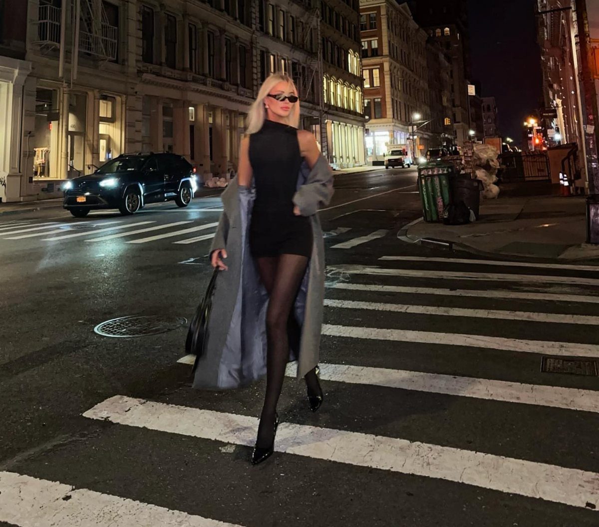 Woman in a black dress and grey coat walking across a city street.