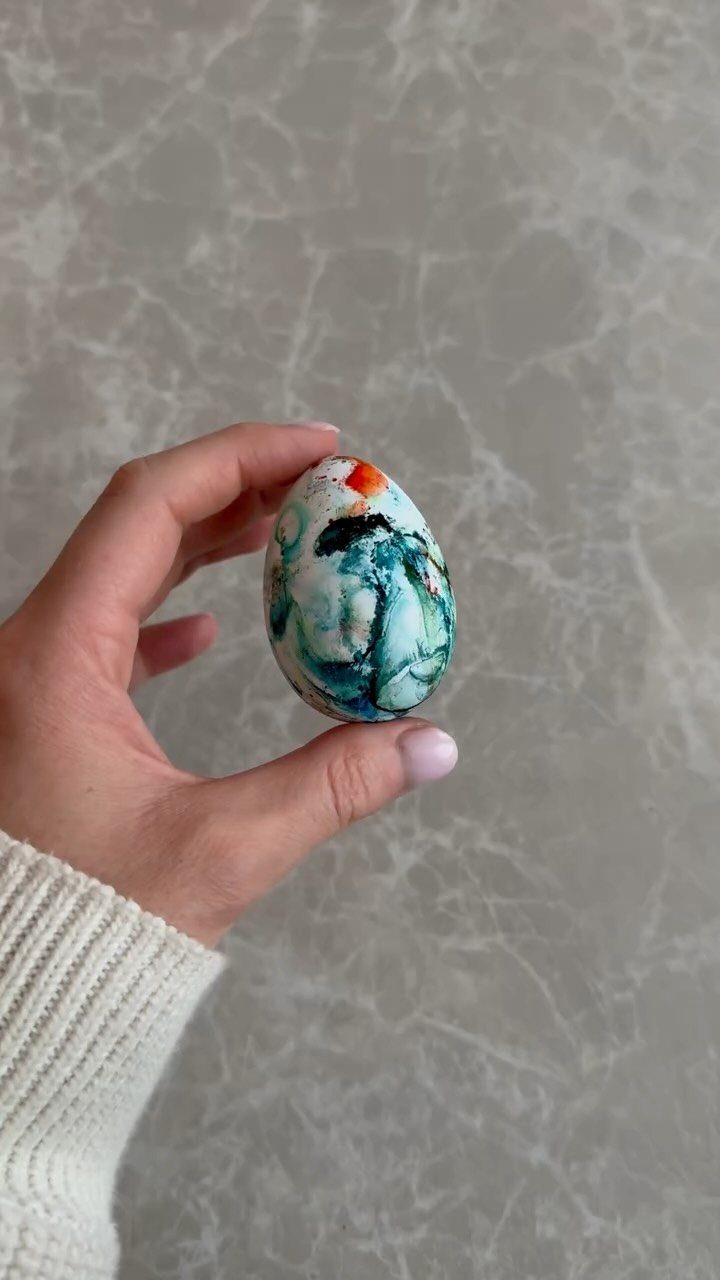 Hand holding a white egg decorated with colorful blue and green paint swirls.